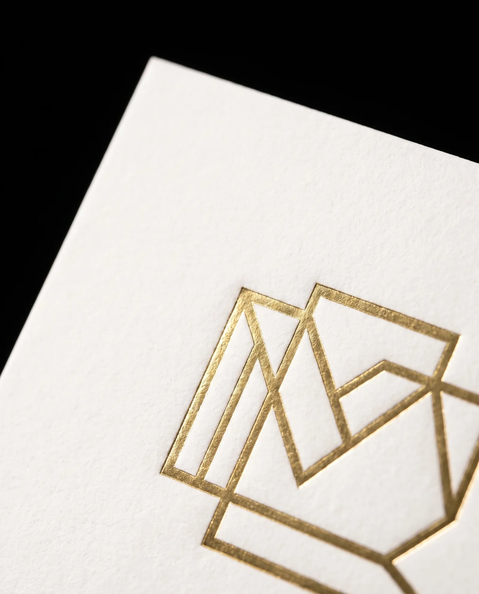 Macro of a gold hot-foil monogram pressed into white cotton, showing the slight bite at the edge of each stroke