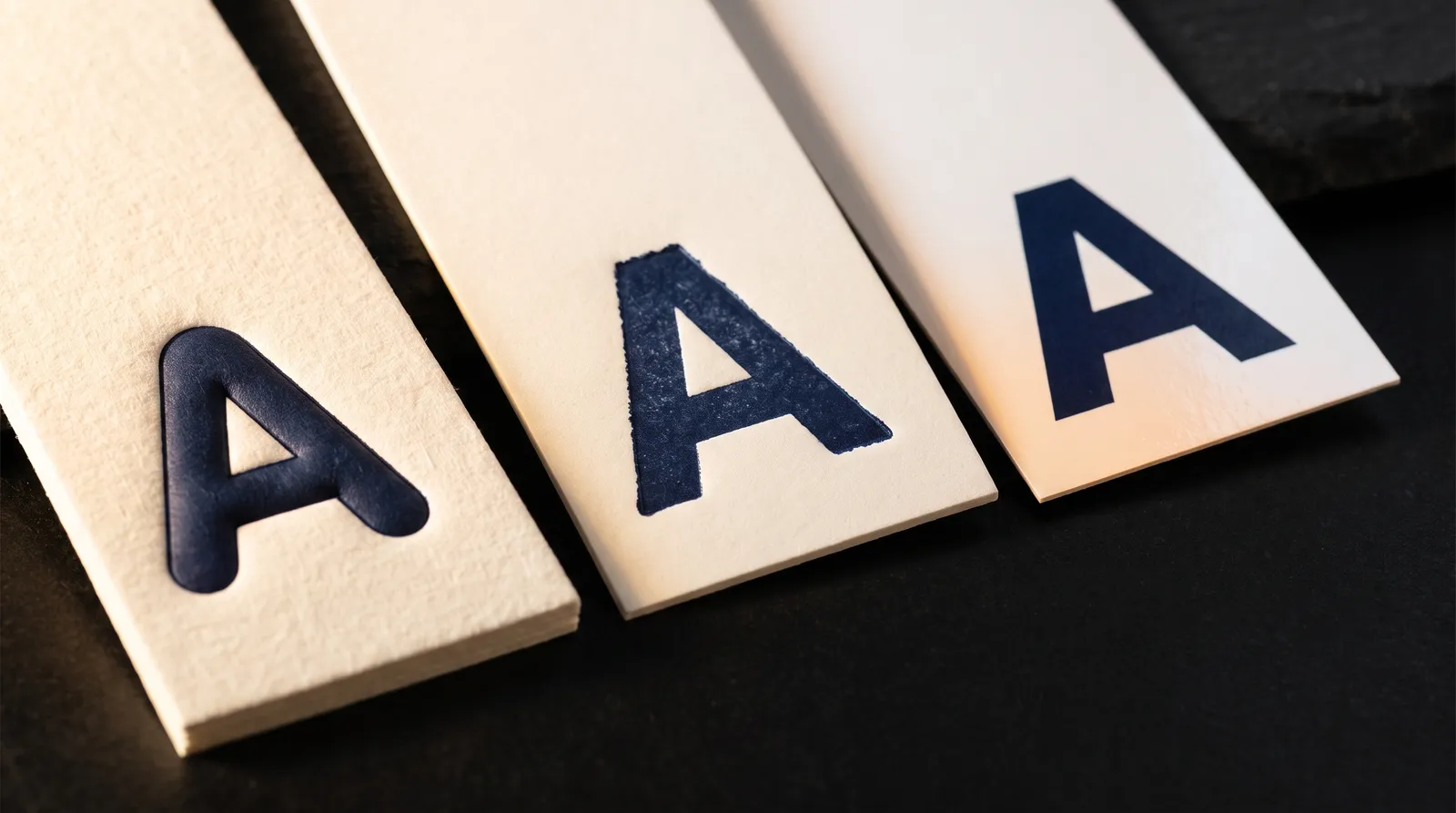 Cotton paper for letterpress compared with wood-pulp and coated paper: the same navy letter pressed deep into cotton, shallower into uncoated wood pulp, and barely marking a coated sheet