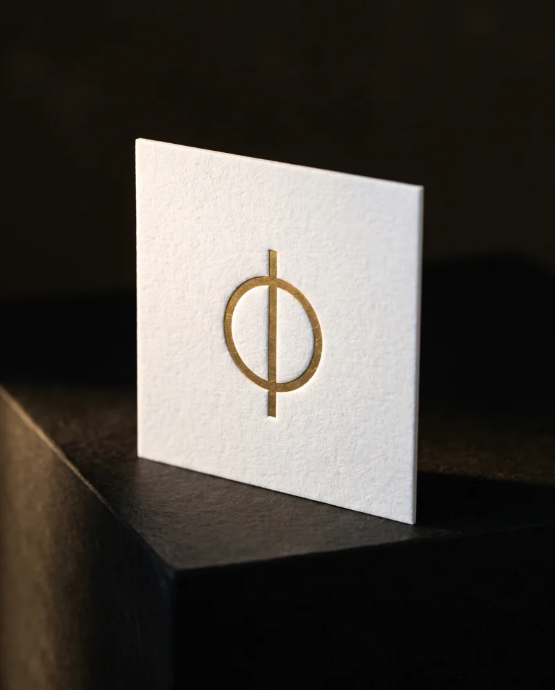 Square 32pt white cotton business card with matte gold foil monogram on black stone