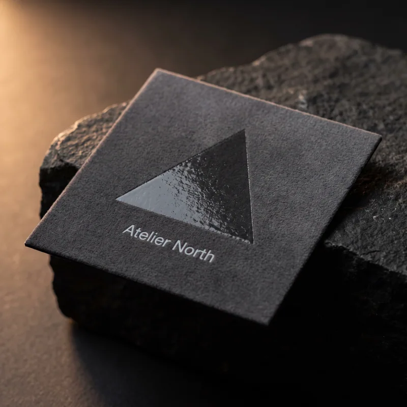 Charcoal soft-touch square business card with clear gloss spot UV triangle, Atelier North